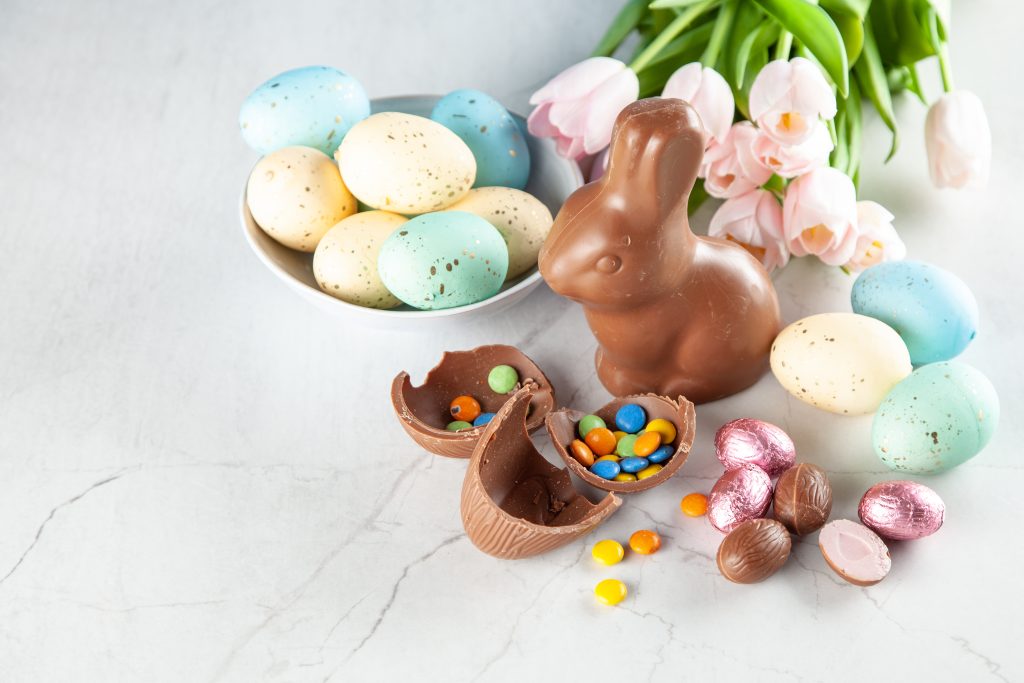 Rechts im Bild ist ein Schokoladen Osterhase zu sehen. Daneben ein Teller mit Ostereiern und vor dem Osterhasen liegen Schokoladeneier.